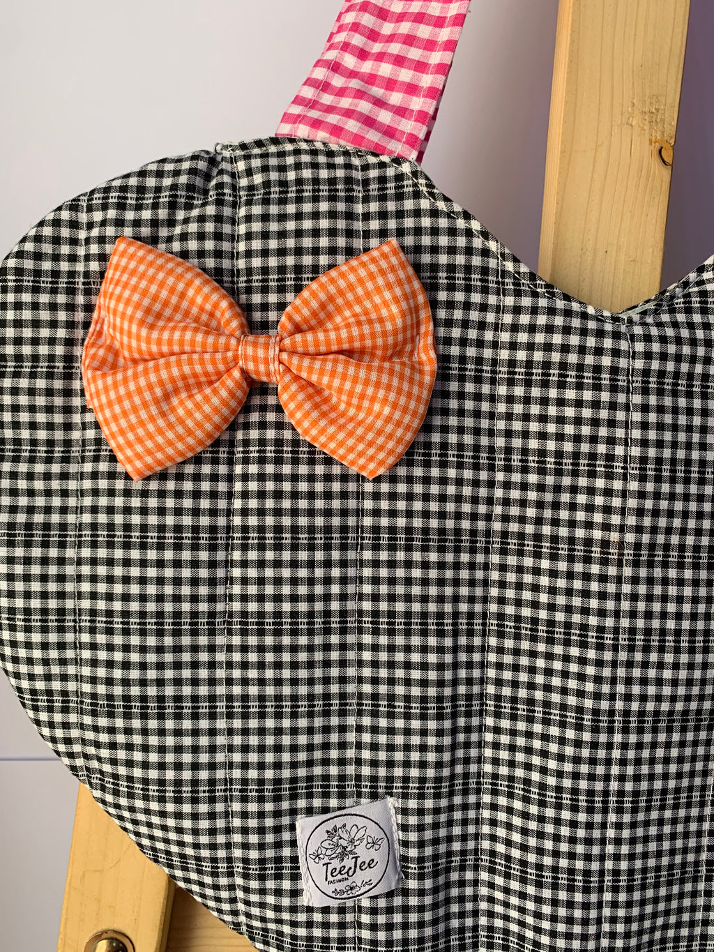 A close-up of a quilted black and white gingham fabric tote bag, featuring a large orange gingham bow on the front and a pink and white gingham strap. A small white sewn label is visible near the bottom.