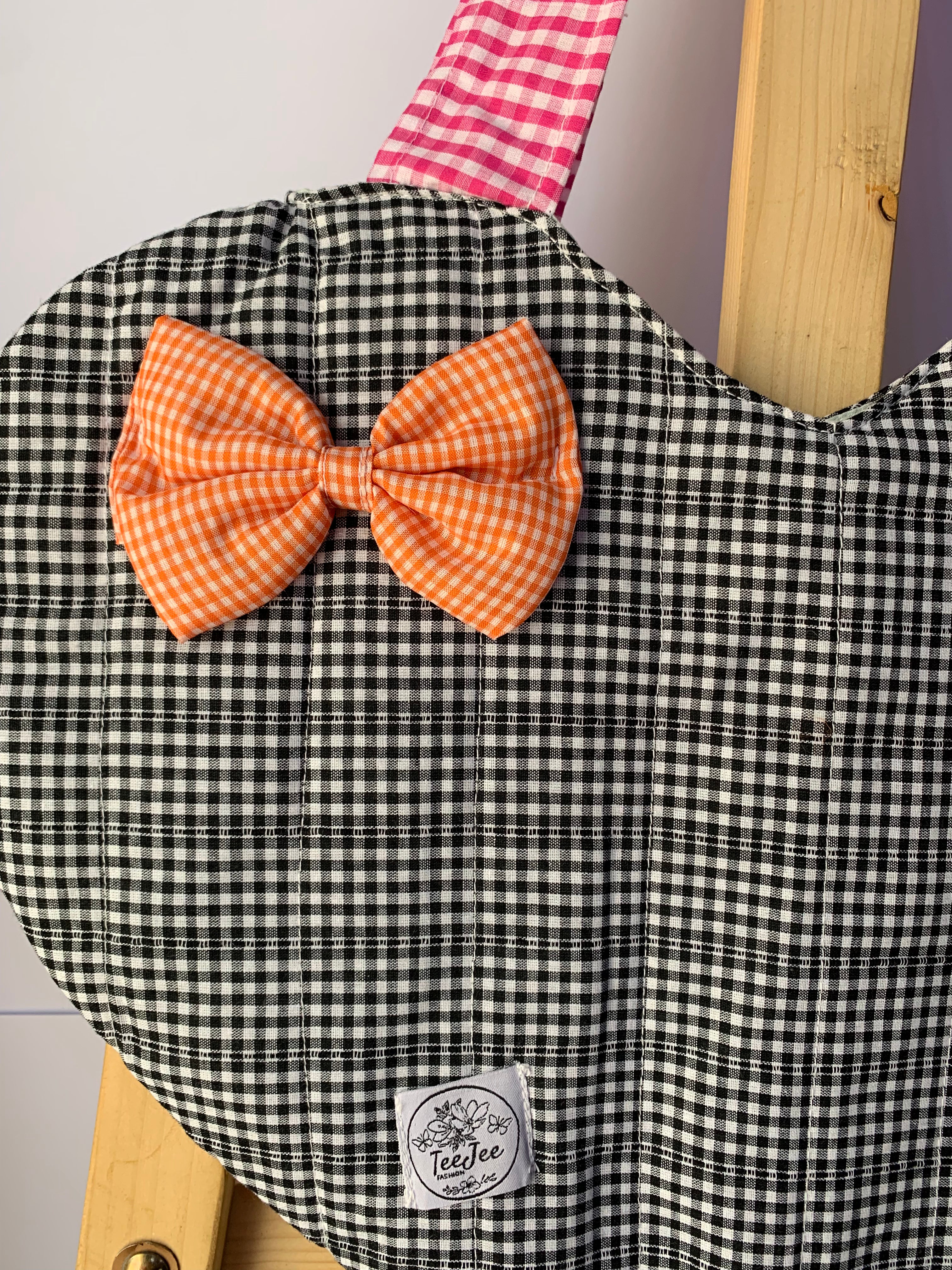 A close-up of a quilted black and white gingham fabric tote bag, featuring a large orange gingham bow on the front and a pink and white gingham strap. A small white sewn label is visible near the bottom.