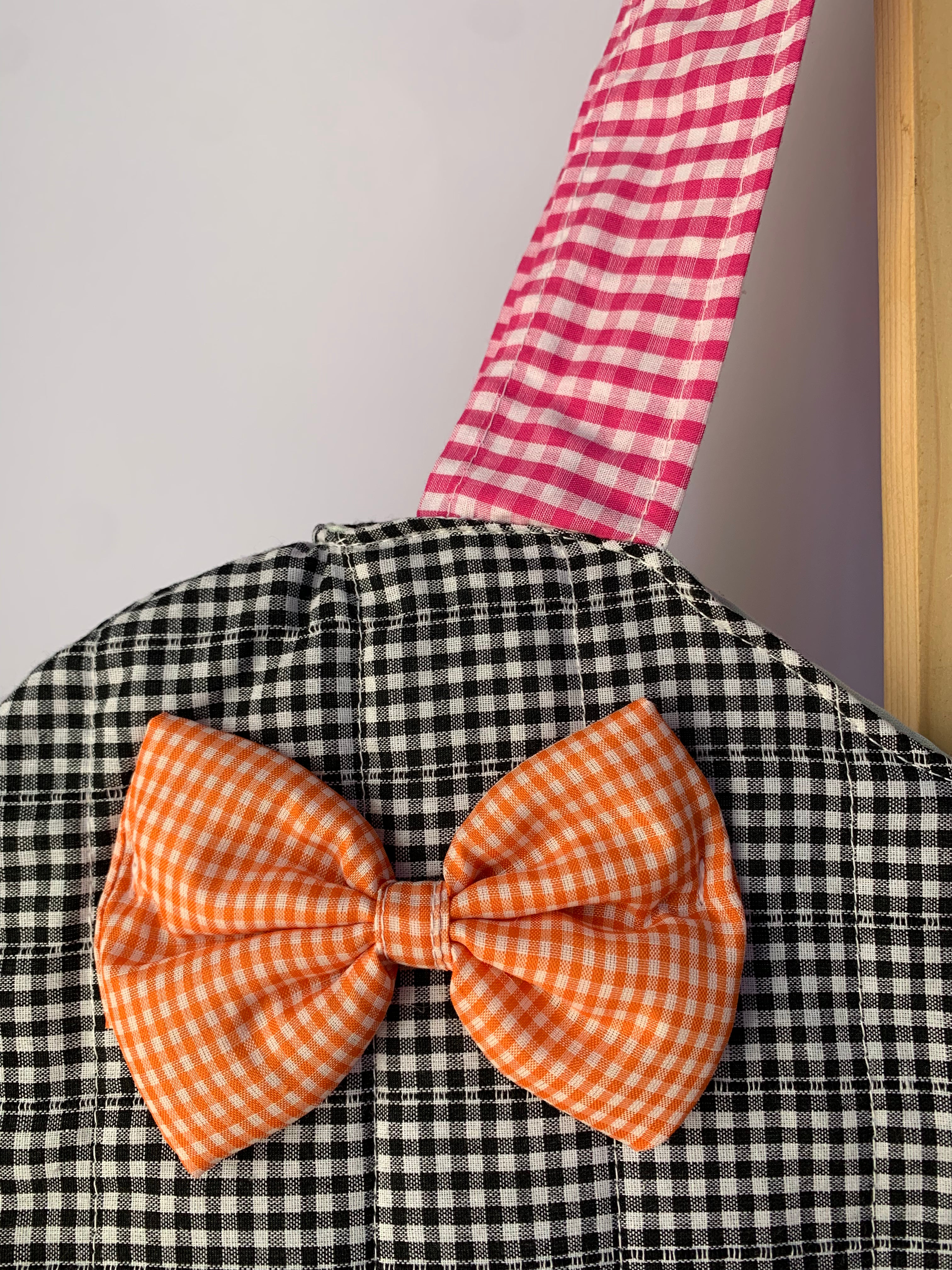 A close-up of a black and white gingham tote bag featuring a prominent orange and white gingham bow on the front, with a pink and white gingham strap hanging from the top against a light gray wall.