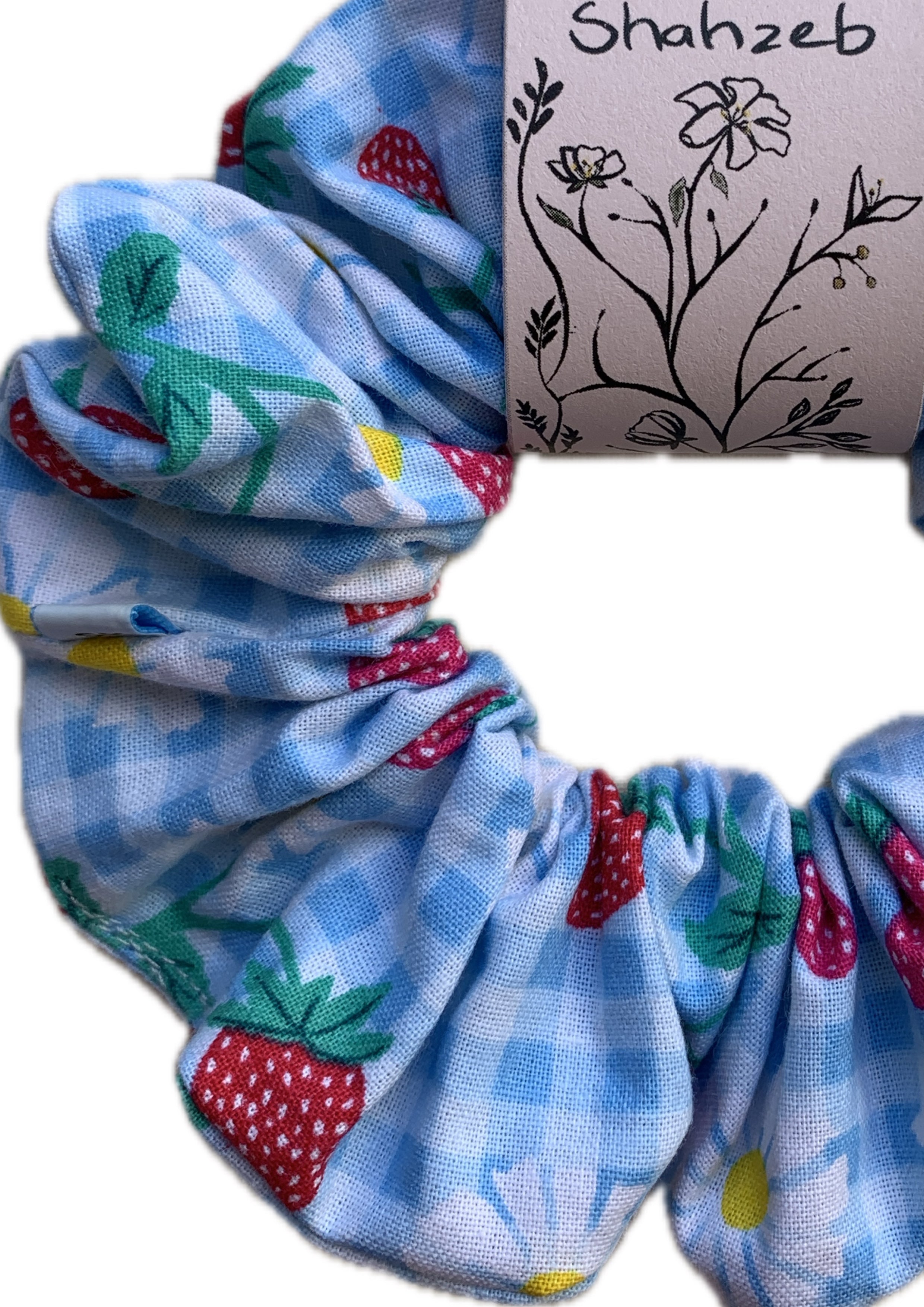A fabric hair scrunchie with a light blue gingham pattern, decorated with colorful strawberry and daisy prints. 