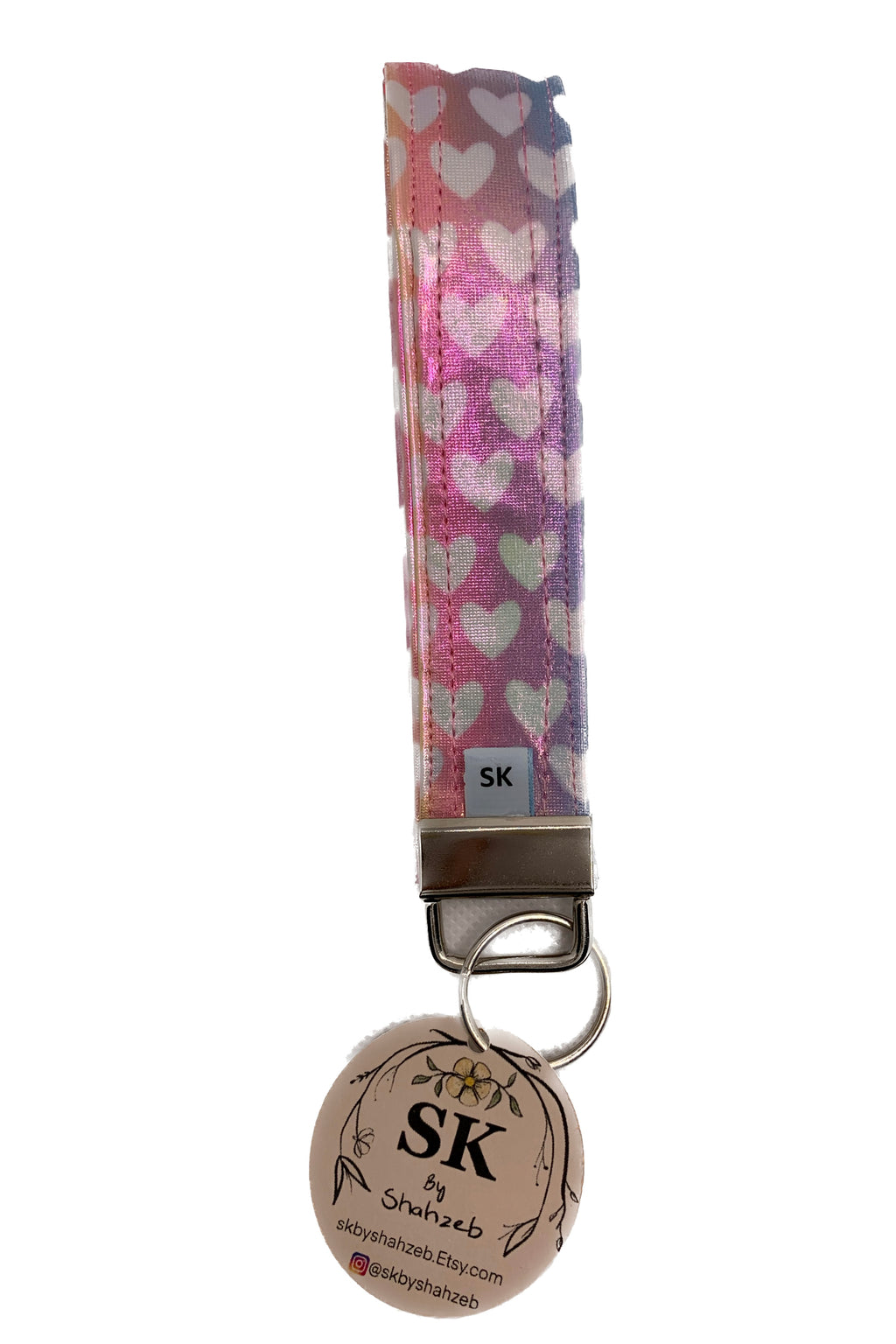 A wristlet strap keychain featuring a pink and purple ombre pattern with white hearts, attached to a silver metal clasp and a round beige tag printed with 'SK' and a floral design.