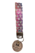 A wristlet strap keychain featuring a pink and purple ombre pattern with white hearts, attached to a silver metal clasp and a round beige tag printed with 'SK' and a floral design.