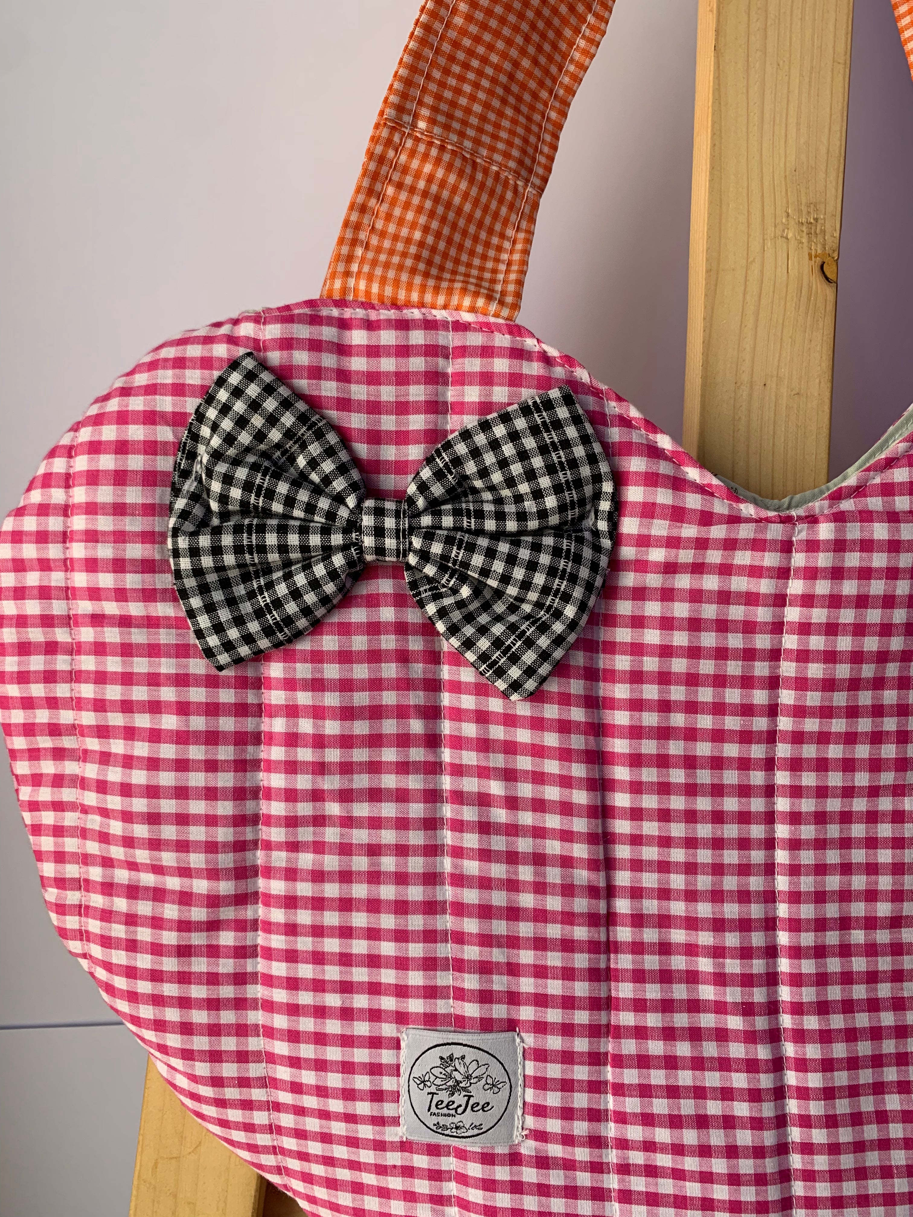 A close-up of a quilted fabric bag with a pink and white gingham pattern, featuring a black and white gingham bow on the front and an orange-brown gingham strap. A small sewn-on label reads 'TeeJee'.