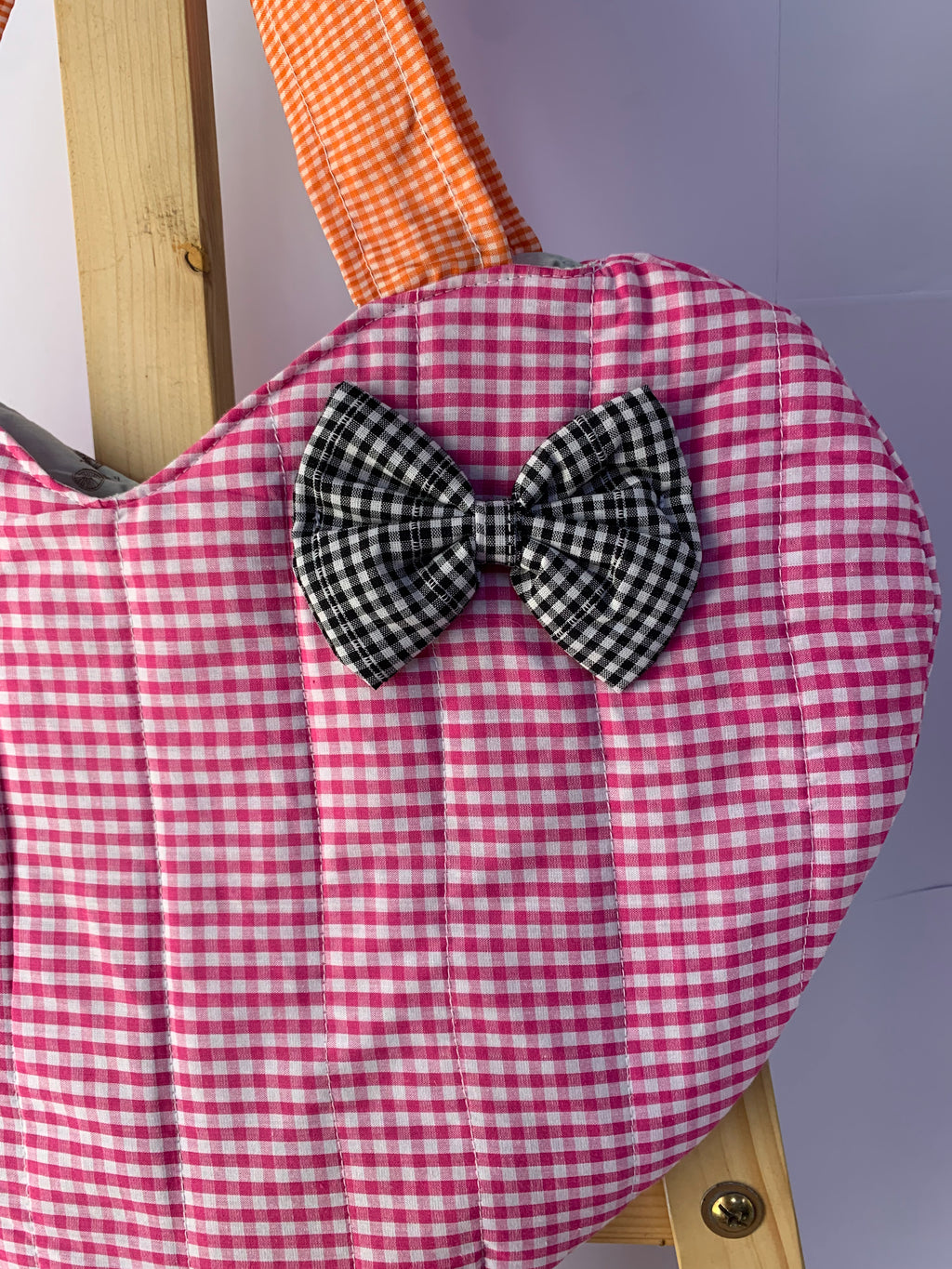 A close-up of a quilted fabric bag with a pink and white gingham pattern, featuring a black and white gingham bow on the front and an orange-brown gingham strap. 