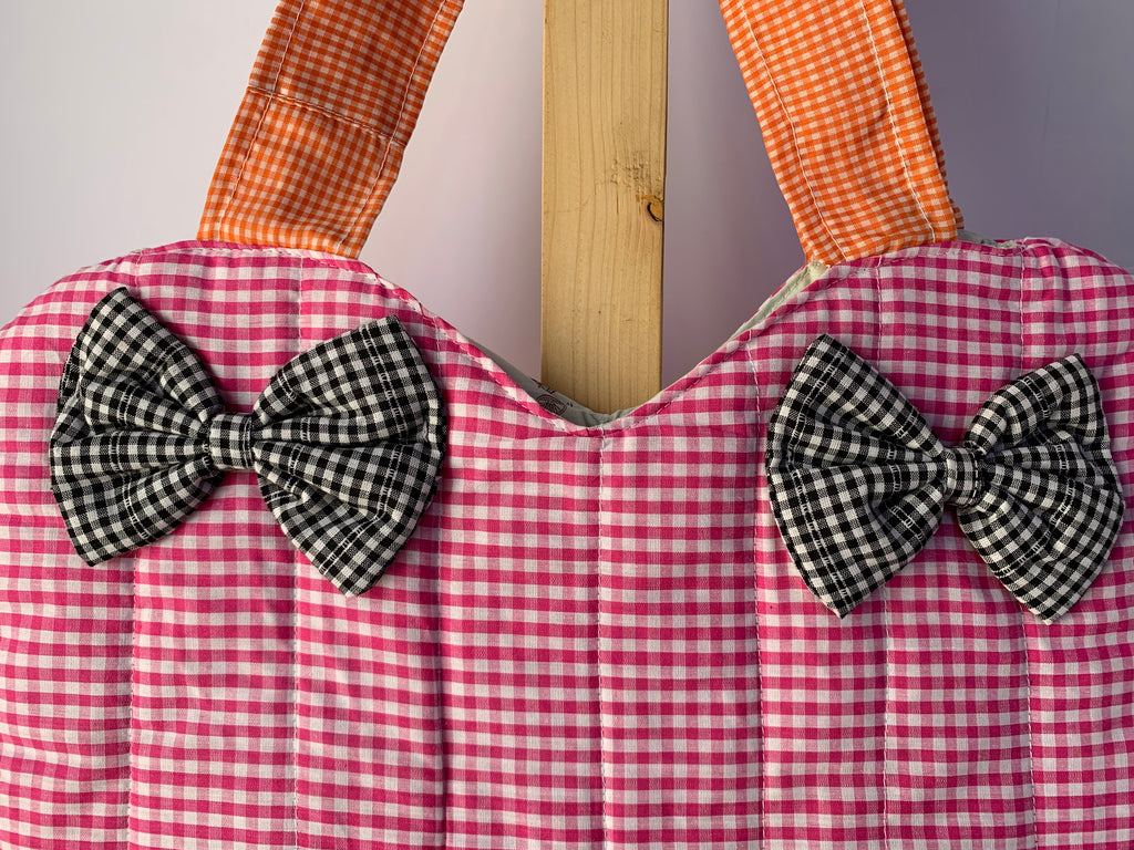 A close-up of a quilted fabric bag with a pink and white gingham pattern, featuring two black and white gingham bow accents on the front and orange-and-white gingham shoulder straps, displayed against a neutral background.