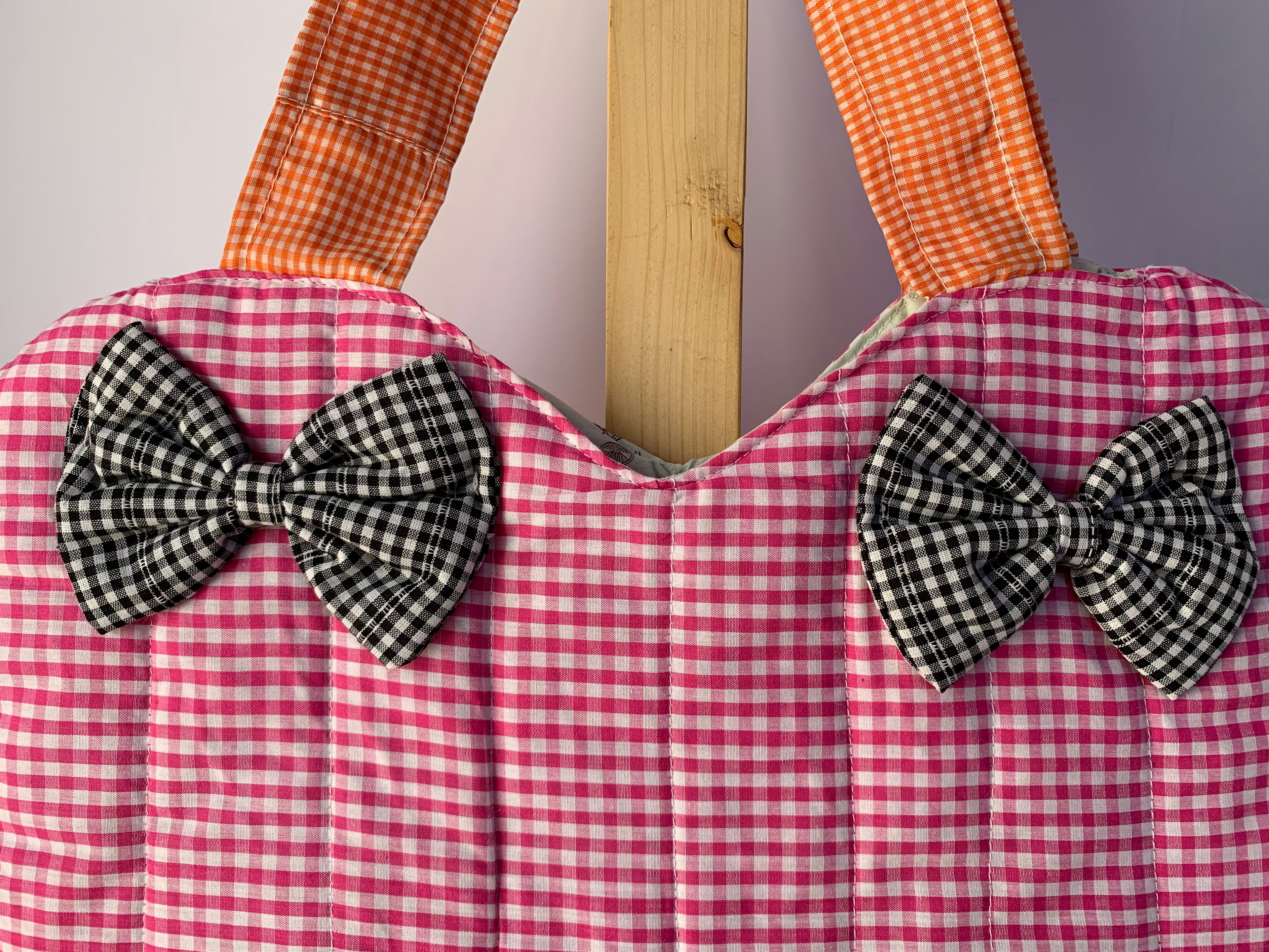 A close-up of a quilted fabric bag with a pink and white gingham pattern, featuring two black and white gingham bow accents on the front and orange-and-white gingham shoulder straps, displayed against a neutral background.