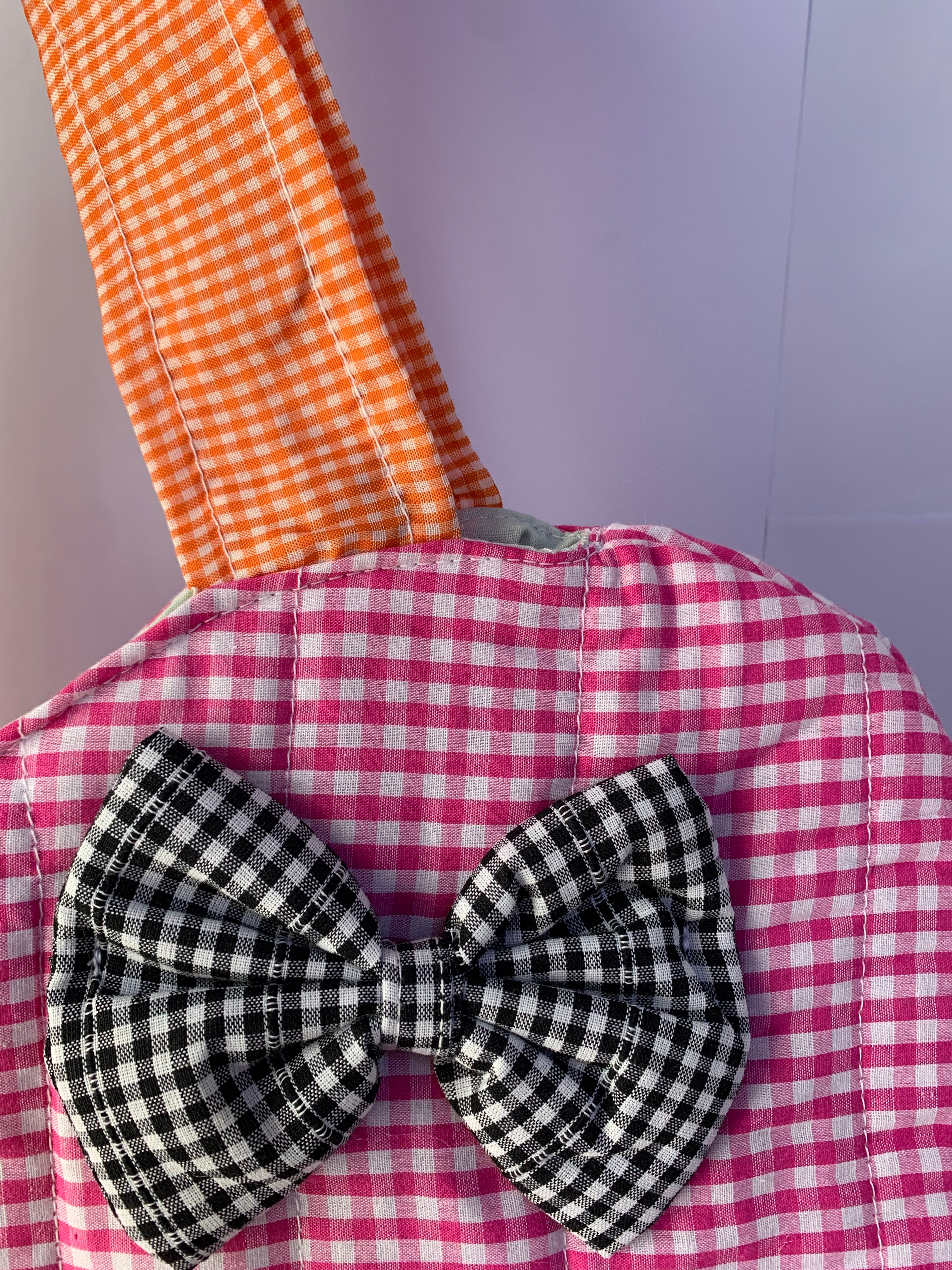 A close-up of a fabric accessory featuring a pink and white gingham body, an orange and white gingham strap, and a large black and white gingham bow in the center.