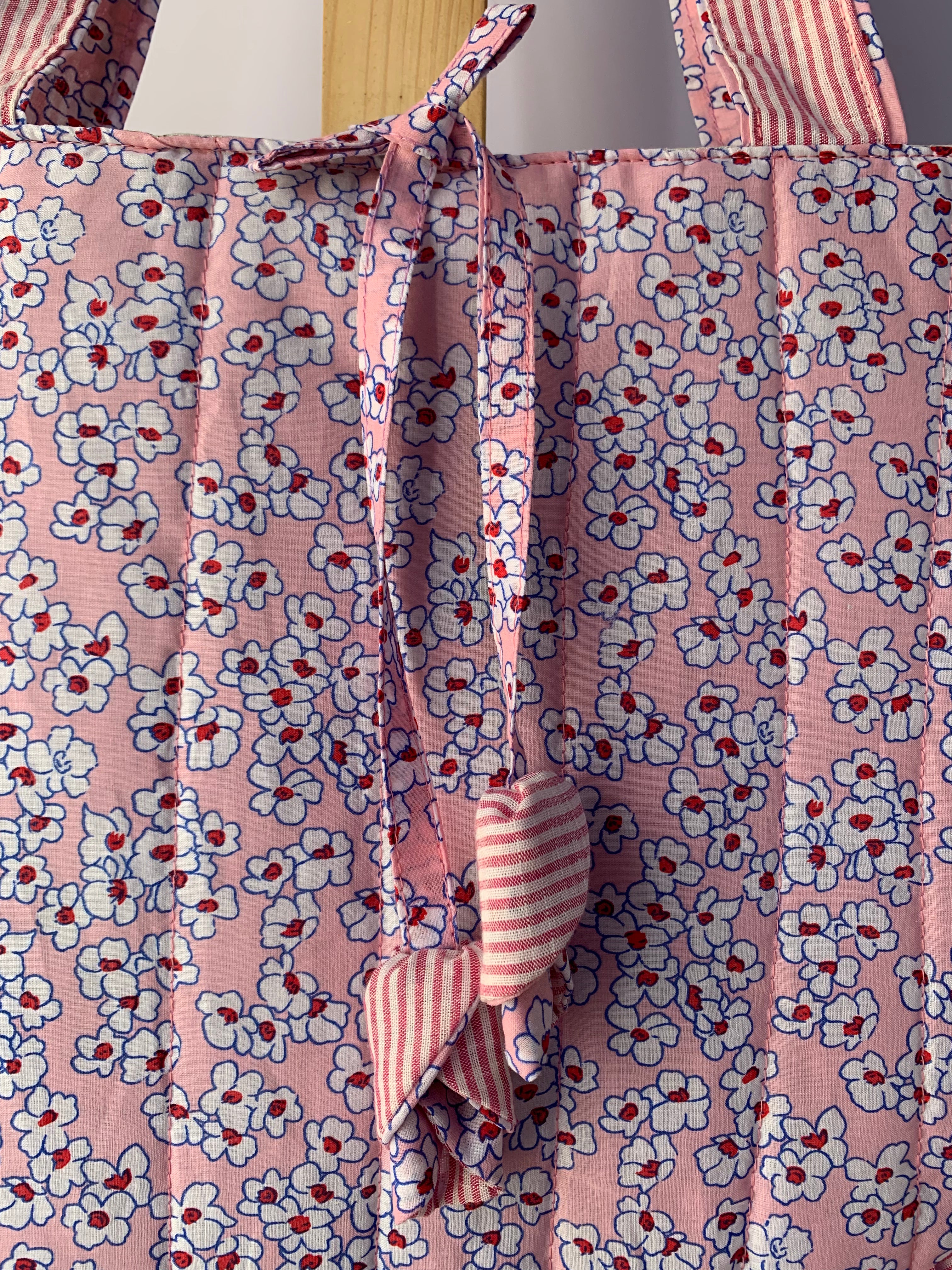 Pink floral tote bag with white and red flowers, featuring a tie closure and striped fabric handles.