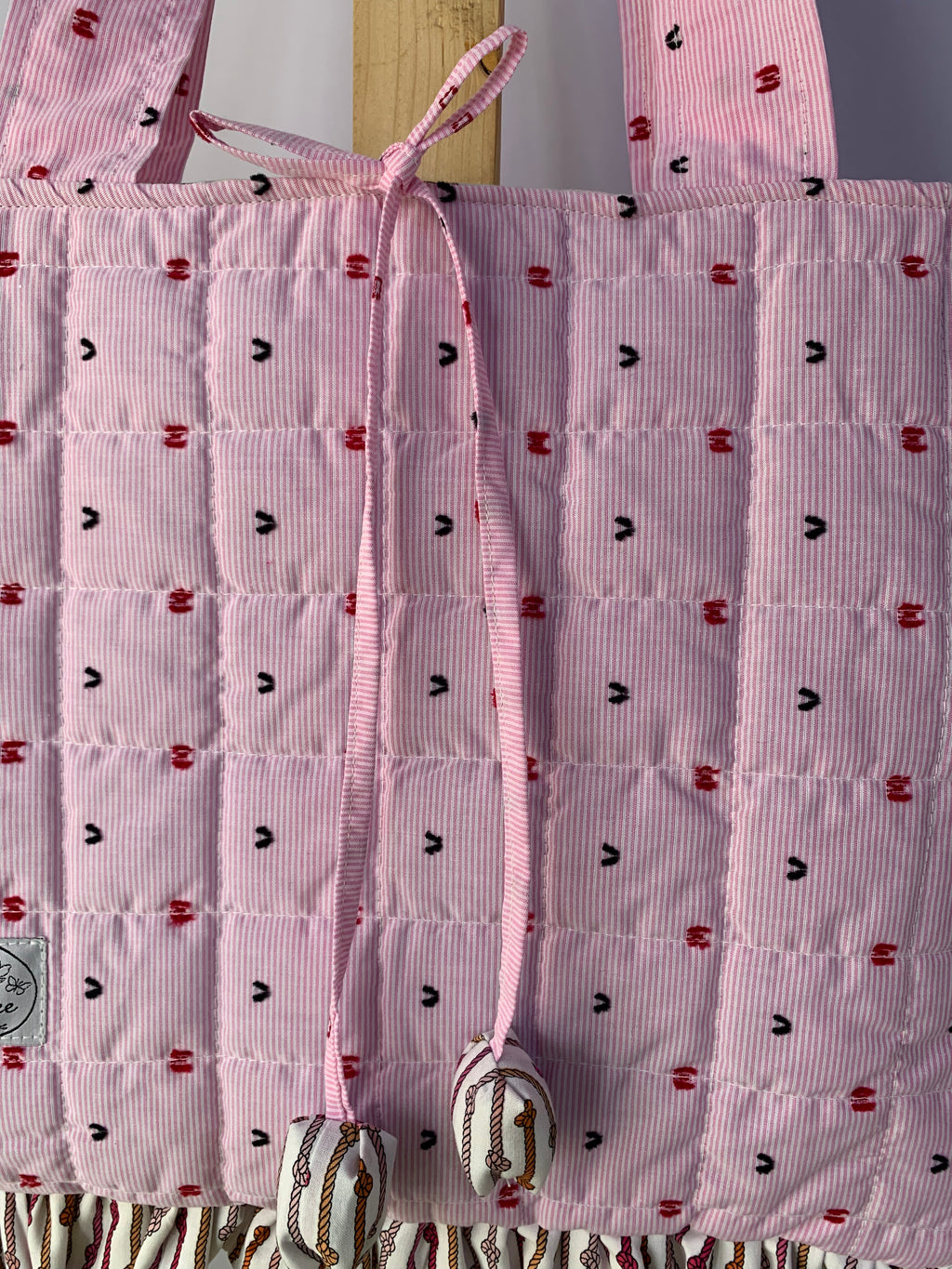 A close-up of a pink quilted  tote bag with a small repeating red and black embroidered pattern, featuring a pink drawstring closure with decorative tassels.
