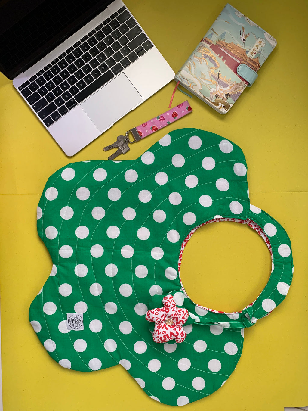 A green and white polka dot tote bag on a yellow surface, styled with a laptop, notebook, and keys nearby.