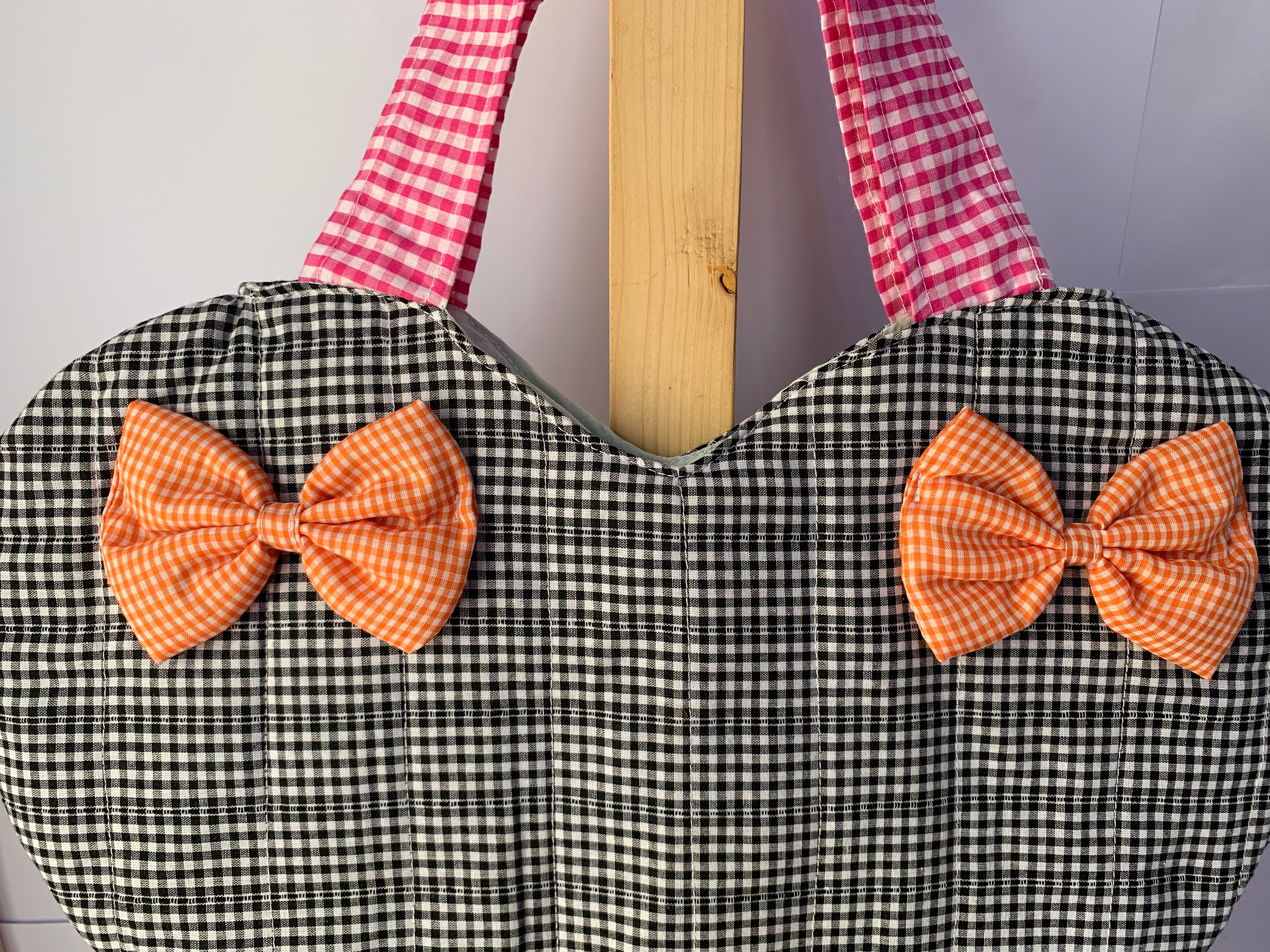 A heart-shaped quilted tote bag with a black and white gingham body, pink and white gingham straps, and two orange gingham fabric bows on the front, displayed on a wooden stand.