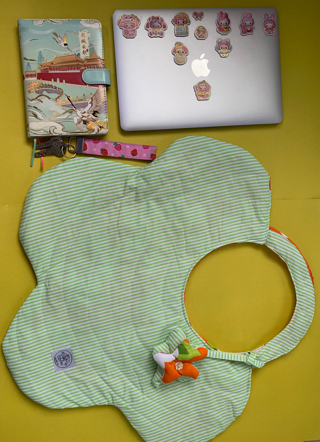 A light green and white striped flower-shaped tote bag with a small orange plush accent, laid flat on a yellow background next to a laptop and a notebook.