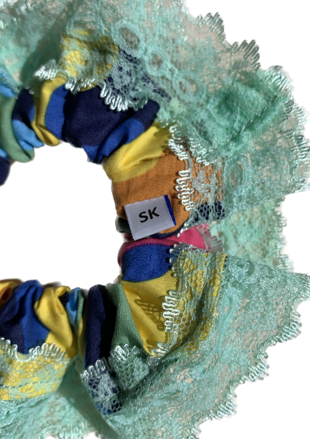 A close-up of a fabric hair scrunchie featuring a ruffled design with a mint green lace trim, multicolour patterned fabric.