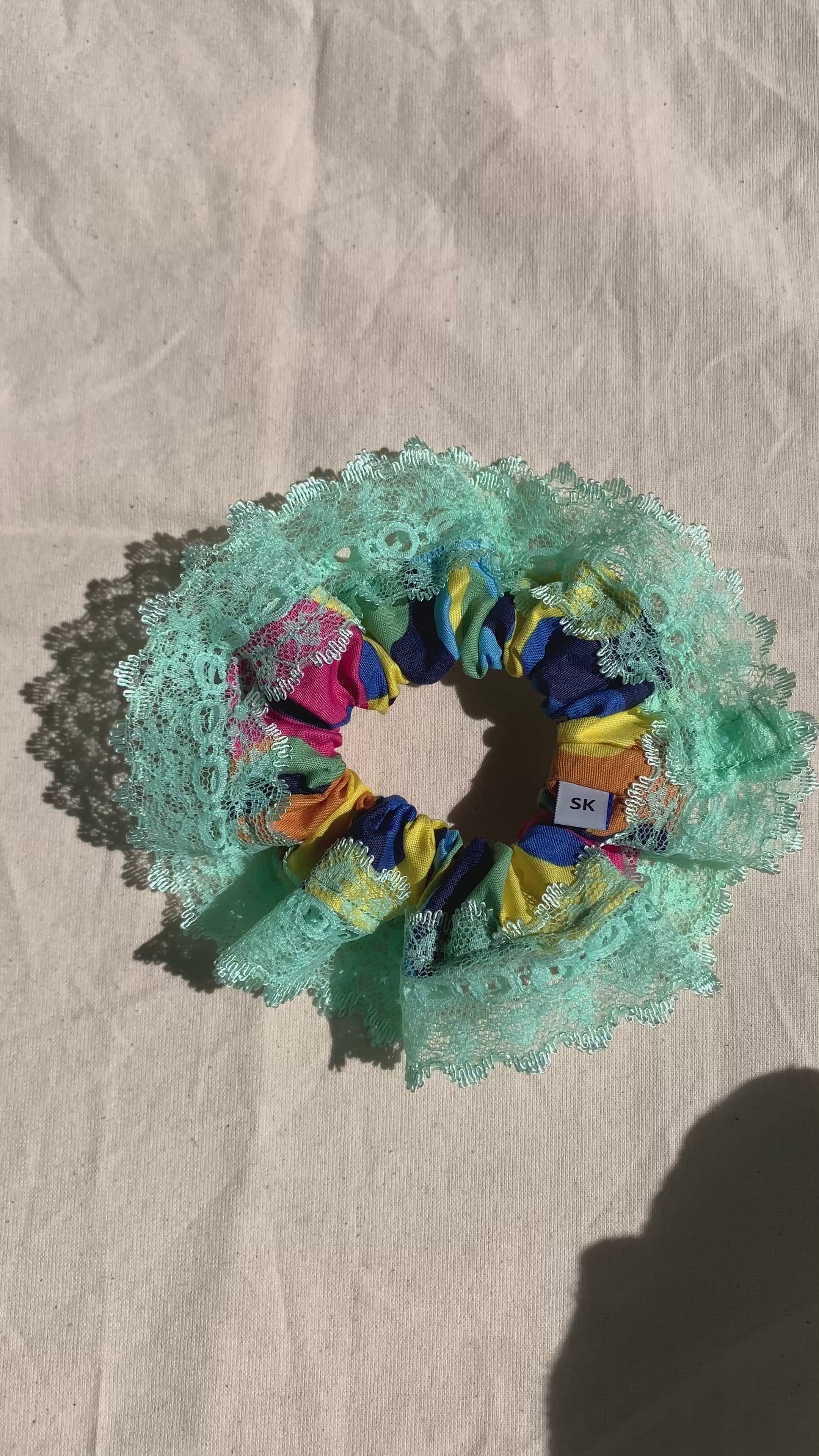 A video showing handmade fabric scrunchie featuring a mint green lace ruffle outer layer and a colorful inner layer with yellow, blue, pink, and orange patterned fabric.