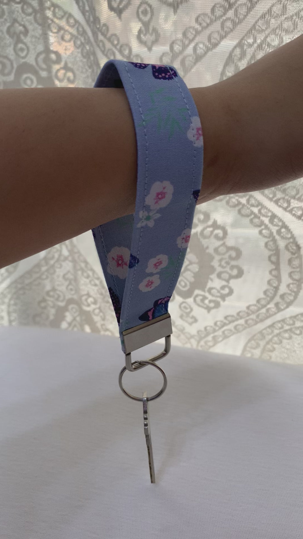 A video showing fabric keychain with a light blue strap featuring a white and pink floral pattern, attached to ring holding a metal key